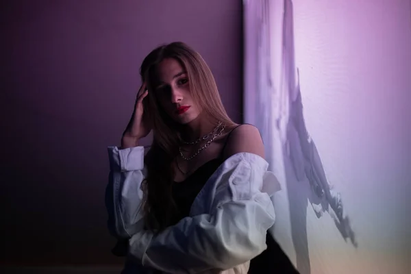 Esmer, gece parıldayan mor bir afişin önünde poz veriyor. Beyaz kot ceketli, siyah tişörtlü, mor ışıkla aydınlatılmış genç bir kız..