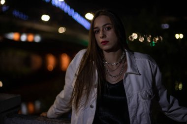 Esmer, gece köprü boyunca parlayan ışıkların arka planında poz veriyor. Bokeh bir kızın arka planında. Geceleri şehirde genç.