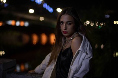 Esmer, gece köprü boyunca parlayan ışıkların arka planında poz veriyor. Bokeh bir kızın arka planında. Geceleri şehirde genç.