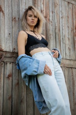 Çıplak omuzlu kot elbiseli bir kız eski ahır kapısına yaslanıyor.