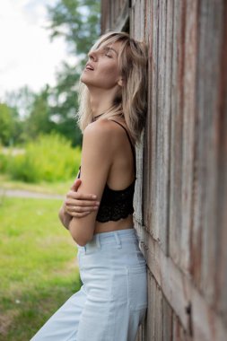 Çıplak omuzlu kot elbiseli bir kız eski ahır kapısına yaslanıyor.