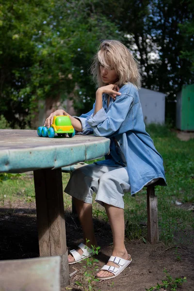 デニムのスーツを着た女の子が庭の外のテーブルのベンチに座り 小さなおもちゃの車を転がしています 大人の子供時代の思い出 — ストック写真