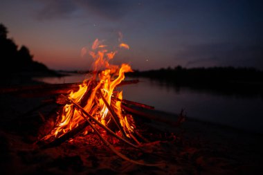 Gece nehir kıyısında şenlik ateşi. Yazın baraj gölünün yanında yanan dallardan ve yakacak odunlardan çıkan kırmızı alevler. Ateşin yanında açık hava eğlencesi..