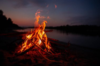 Gece nehir kıyısında şenlik ateşi. Yazın baraj gölünün yanında yanan dallardan ve yakacak odunlardan çıkan kırmızı alevler. Ateşin yanında açık hava eğlencesi..