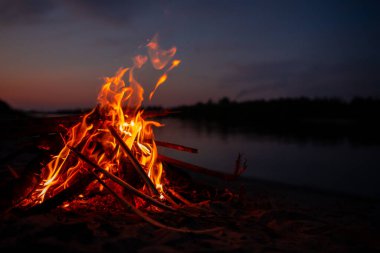 Gece nehir kıyısında şenlik ateşi. Yazın baraj gölünün yanında yanan dallardan ve yakacak odunlardan çıkan kırmızı alevler. Ateşin yanında açık hava eğlencesi..