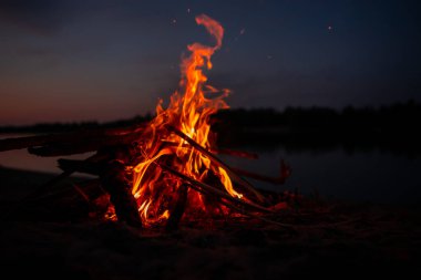 Gece nehir kıyısında şenlik ateşi. Yazın baraj gölünün yanında yanan dallardan ve yakacak odunlardan çıkan kırmızı alevler. Ateşin yanında açık hava eğlencesi..
