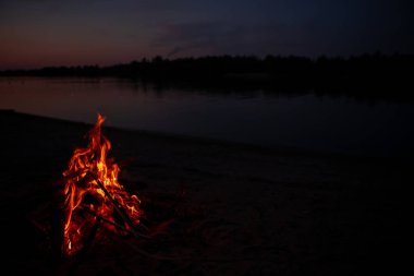 Gece nehir kıyısında şenlik ateşi. Yazın baraj gölünün yanında yanan dallardan ve yakacak odunlardan çıkan kırmızı alevler. Ateşin yanında açık hava eğlencesi..