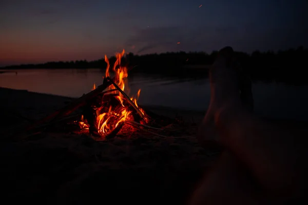 男子的脚在河岸靠近火边的夜晚 火光照亮了一个人的脚 夏天在水库附近燃烧的树枝和木柴燃起的红色火焰 待在新鲜空气里 — 图库照片