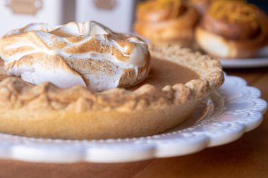 Süslü balkabağı turtası beyaz bir tabağın üstünde kremayla süslenmiş ve arka planda tarçın ruloları var..