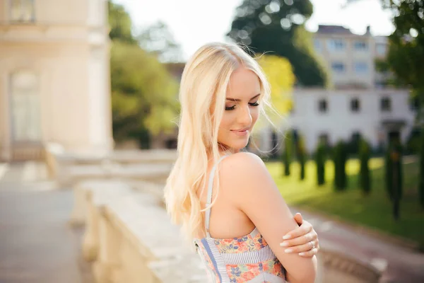 Stilvolle blonde Frau, die im Sommer bei Sonnenuntergang draußen auf der Straße in der Stadt spaziert und ein Kleid mit nacktem Rücken trägt. Rückansicht. — Stockfoto