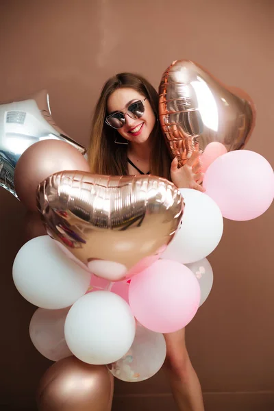 Slank meisje in een bodysuit en gekleurde ballonnen in haar handen staat op een bruine achtergrond in een feestelijke stemming poseren voor de camera. — Stockfoto