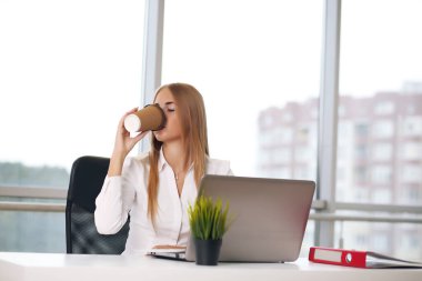 Genç ve güzel bir iş kadını parlak modern ofiste dizüstü bilgisayarda çalışıyor, pencereden dışarı bakıyor.