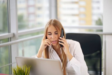 Genç ve güzel bir iş kadını parlak modern ofiste dizüstü bilgisayarda çalışıyor, pencereden dışarı bakıyor.