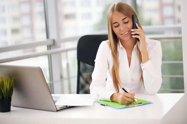 Jovem mulher bonita alegre falando no telefone e olhando para a câmera com um sorriso sentado em seu local de trabalho — Fotografia de Stock