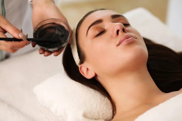 Closeup of unrecognizable cosmetologist applying face mask with brush to face of beautiful woman lying on massage table in SPA center — Stock Photo, Image