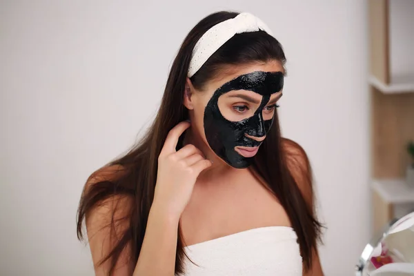Młoda kobieta z czarną maską detoksykacyjną na twarzy, nastolatka dba o tłustą skórę, oczyszczając pory. Salon piękności. Skóra. — Zdjęcie stockowe