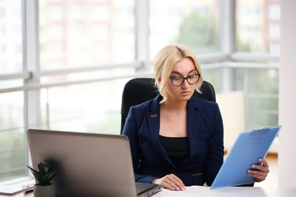 Zdjęcie poważnej kierowniczki lub reżyserki noszącej formalną odzież i okulary trzymające w ręku dokumenty papierowe podczas pracy w biurze na laptopie — Zdjęcie stockowe