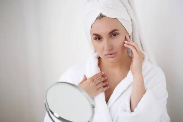 Wanita muda dengan kulit sempurna, menerapkan krim pelembab di wajahnya. Foto wanita setelah mandi dalam jubah mandi putih dan handuk di latar belakang putih. Konsep perawatan kulit — Stok Foto