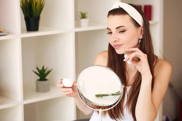 Aantrekkelijke jonge vrouw in badkamer na douche staat voor de spiegel met gezichtscrème. Vrouwen geven om. — Stockfoto