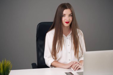 Gri bir ofiste dizüstü bilgisayarla masada oturan genç bir kadın. öğrenci ya da iş kadını