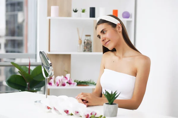 Jeune fille brune prend soin de sa peau, debout devant un miroir, profitant de soins de beauté pour elle-même, souriant tendrement Photos De Stock Libres De Droits