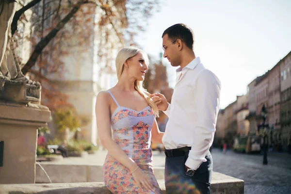 Hermosa pareja caminando por la cuadra. Hombre de pelo oscuro con una camisa blanca abrazando a una rubia en un hermoso vestido —  Fotos de Stock