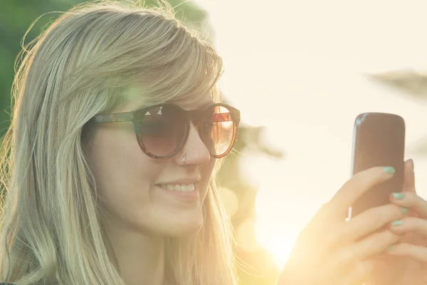 woman in sunglasses typing on smartphone while standing on blurred natural background