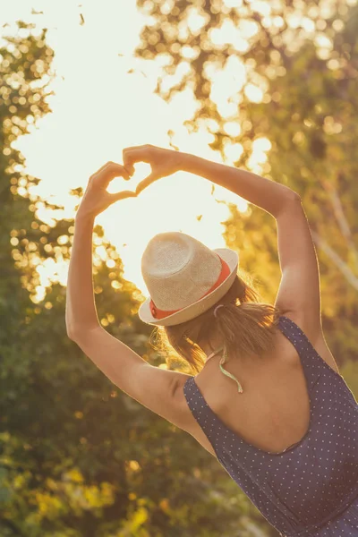 arkadan görünüşü şapka giyen kadın ve elbise kaldırdı elleriyle ayakta ve kalp işareti ormanda güneş doğarken gösterilen.