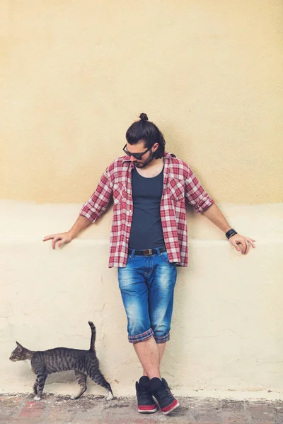handsome man with hairstyle in sunglasses and trendy clothes posing with cat on beige wall background