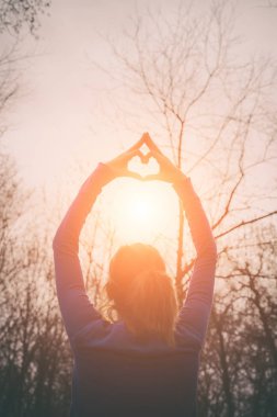 arkadan görünüşü kaldırdı elleriyle ayakta ve kalp işareti ormanda güneş doğarken gösterilen kadın