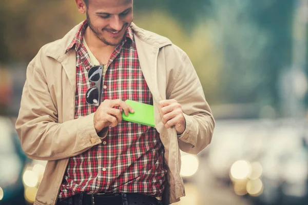 smiling man wearing stylish clothes taking out green smartphone of pocket while walking at street