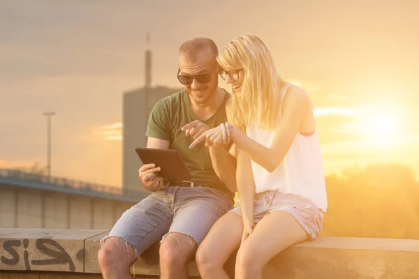 sweet couple using tablet pc while sitting on roof at sunset with cityscape on background