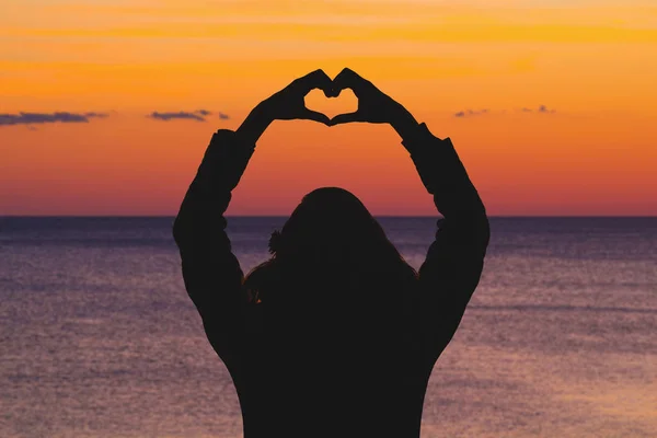 back view of woman with raised hands showing heart sign while standing at seashore at orange fiery sunset
