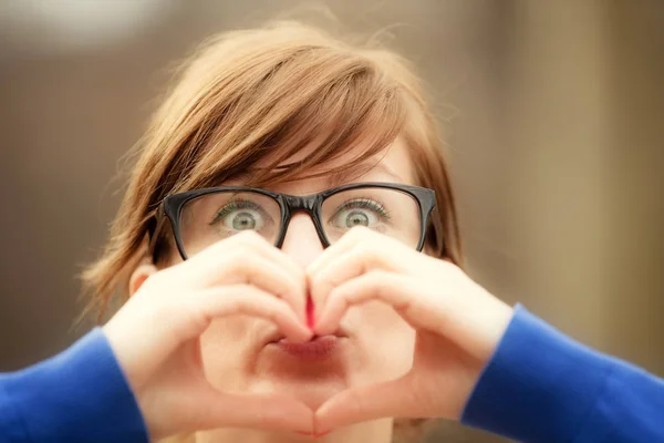 cute woman with big eyes looking at camera and showing heart sign on blurred natural background , close-up
