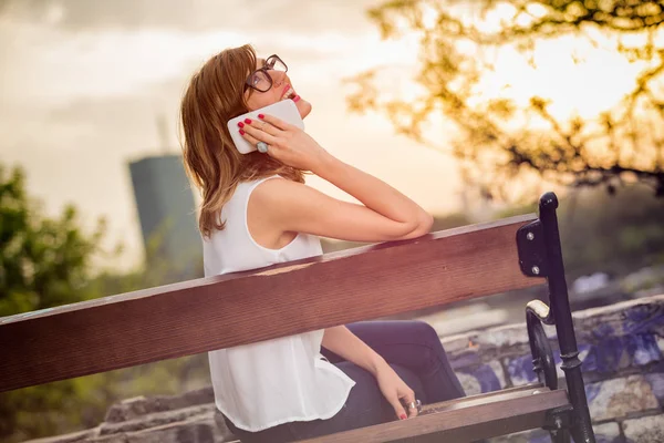 happy woman wearing summer clothes talking on smartphone while sitting on bench at street