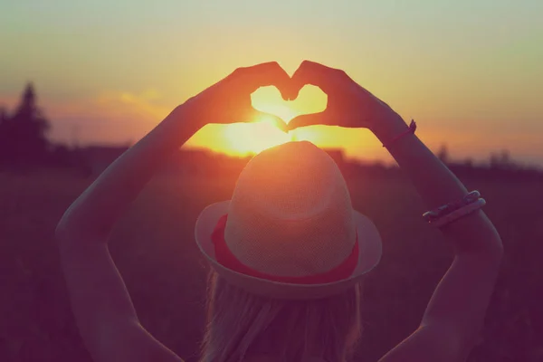 back view of woman in hat with raised hands showing heart sign at sunset on sky background