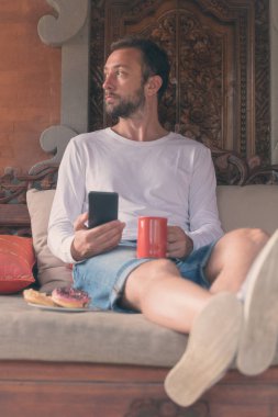 Cep telefonu, kahve kupa ile donats kanepede tutan adam.