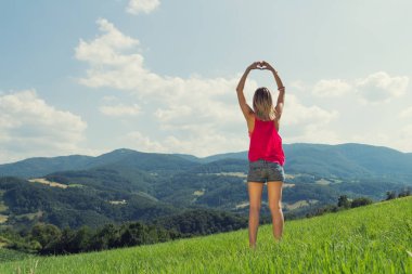 Kız arka planda dağ manzarası olan bir kalp-şekil yapma.