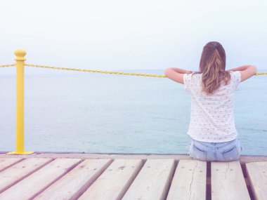 Ahşap bir köstebek üzerinde oturan kız / iskele ve okyanusa bakarak.