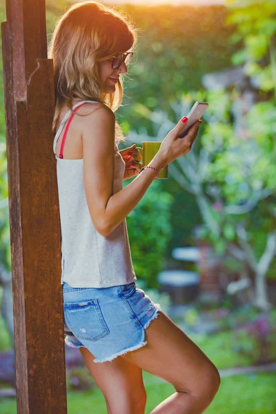 Młoda Kobieta Przy Użyciu Telefonu Komórkowego Picia Kawy Herbaty Werandzie — Zdjęcie stockowe