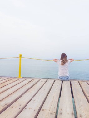 Ahşap bir köstebek üzerinde oturan kız / iskele ve okyanusa bakarak.