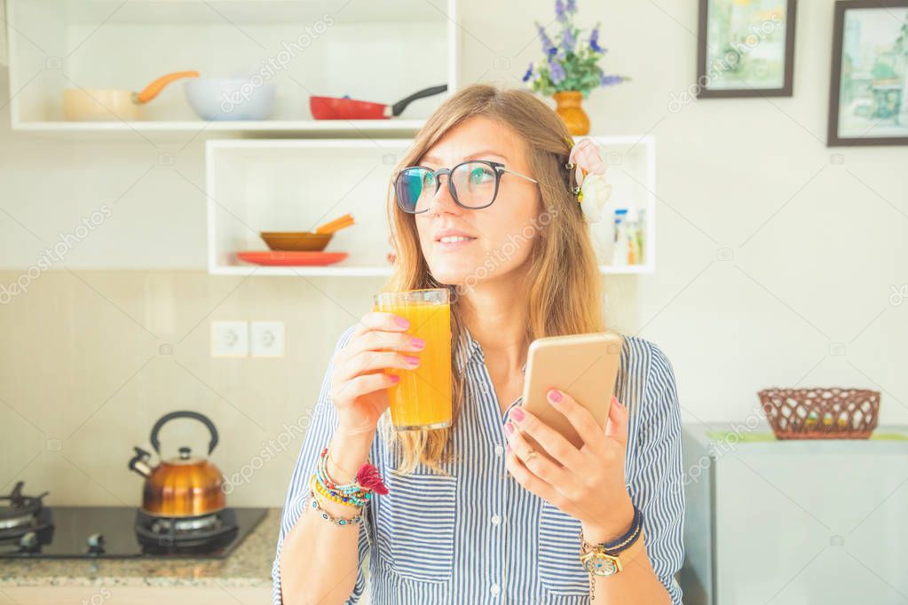 Cute girl drinking fresh orange juice and using cellphone in the kitchen.