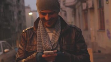 Genç adam akıllı telefon ve şehirde gülümseyen deri ceket ve şapka manifatura ileti