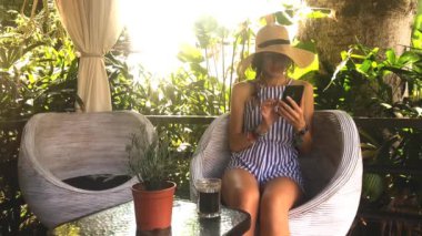 Genç kadın elbise ve şapka manifatura smartphone kullanarak ve koltukta oturup çay içme arkadaşlar