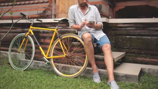 若い男が自転車のそばに座ってアジアの庭でスマート フォンを使用して — ストック動画