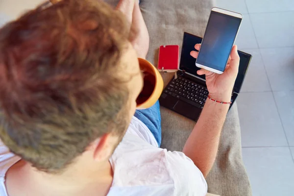 Homem usando celular e laptop enquanto bebe café / chá no th — Fotografia de Stock