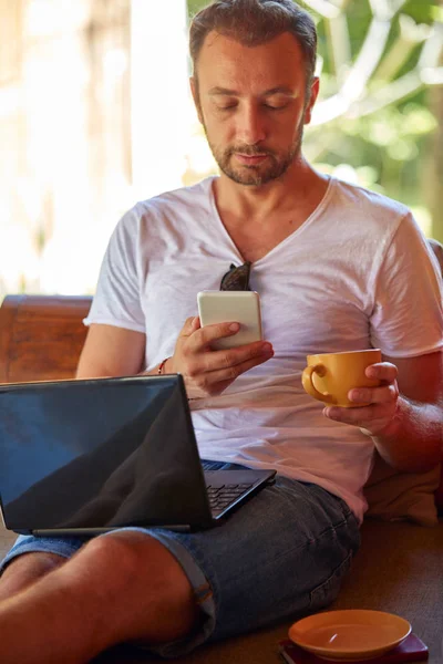 Hombre usando el teléfono celular y el ordenador portátil mientras bebe café / té en th — Foto de Stock