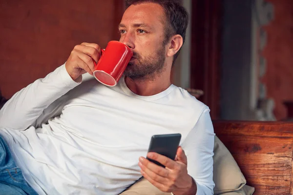 Hombre sentado en un sofá terraza y beber café / té mientras usa — Foto de Stock