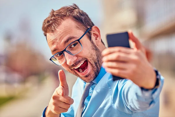 Divertido hombre de negocios con pulgares hacia arriba mientras usa el teléfono inteligente al aire libre —  Fotos de Stock