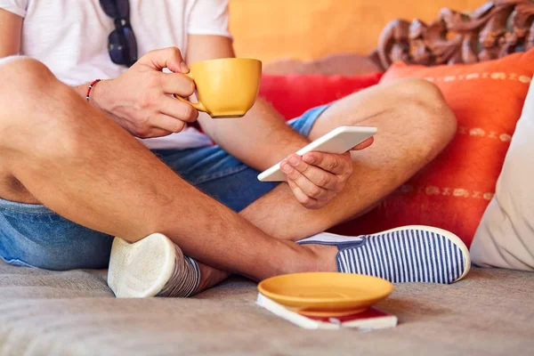 Man met behulp van mobiele telefoon tijdens het drinken van koffie/thee op de Bank. — Stockfoto